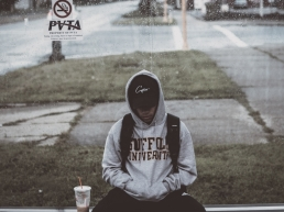 African American teenager, being patient waiting on the bus wearing a hooded sweatshirt, sweatpants and a baseball cap with a drink on his right
