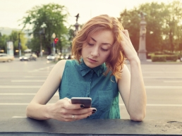 Caucasian woman with red hair, sitting on a street corner look at her online dating app on her cell phone frustrated