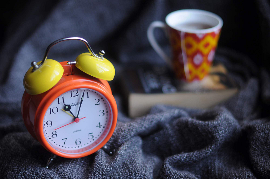 Red-Orange clock with yellow bells, a book and a red and yellow cup, laying on top of a navy blanket