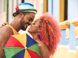 Teen male with hat and beard, celebrating a date a Carnival with a female with orange curly hair and a multicolored umbrella