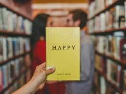 Happiness image of a man and woman in a library aisle facing each other. Another person whose arm is visible, is holding a yellow book titled, 
