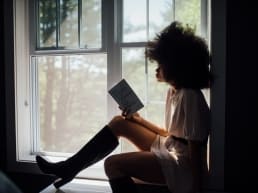 tough girls: African American woman with an afro in a window reading a book.