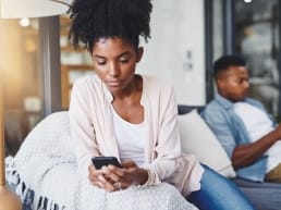 African American woman with curly hair, and man in communication via text message on their cell phones while sitting on opposite ends of the couch