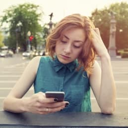Caucasian woman with red hair, sitting on a street corner look at her online dating app on her cell phone frustrated